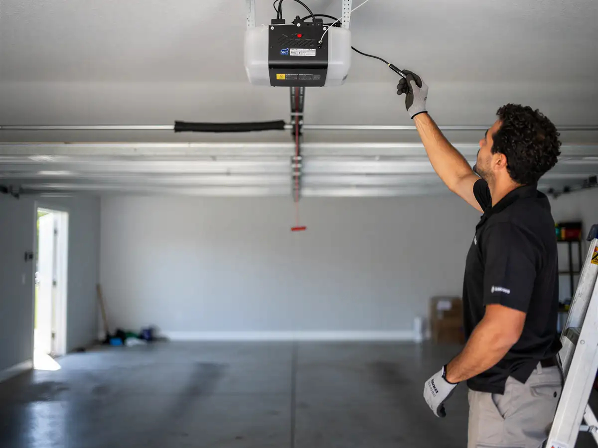 A technician on a ladder adjusts a garage door opener in an empty garage.